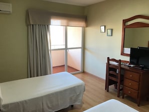 Habitación de hotel con cama, escritorio con ordenador, silla, cómoda y puerta corrediza de vidrio que da a un balcón.
