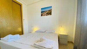 A neatly made bed with white linens and towels, a wooden door, a small bedside table with a lamp, and a framed picture on the wall. - Las Gondolas Apartments (San Javier)