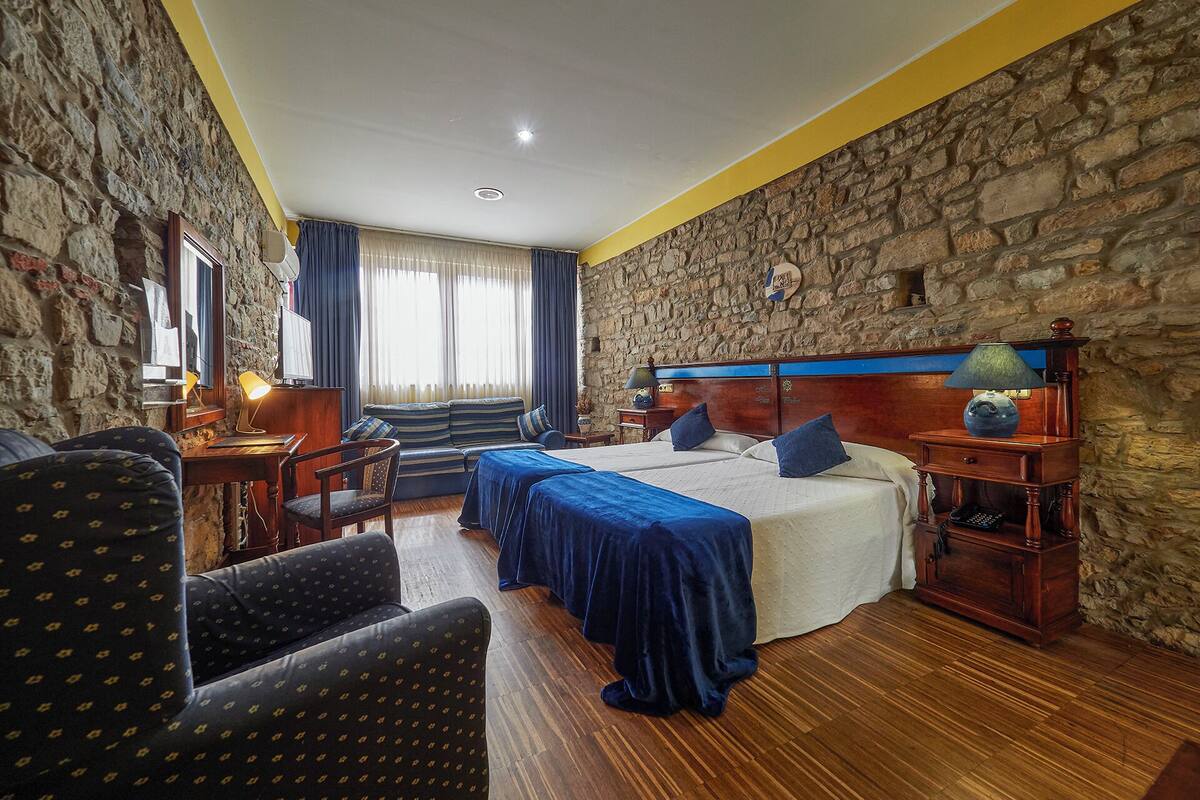 Habitación de hotel con pared de piedra, cama con ropa de cama azul, escritorio con lámpara y una silla.