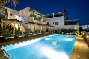 A swimming pool with lounge chairs and umbrellas, surrounded by buildings with balconies and a staircase. - Hotel Andreas (Santorini)