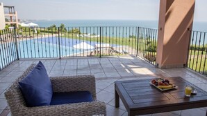 A balcony with a wicker sofa, a wooden table with fruit and juice, and a view of the sea and pool.
