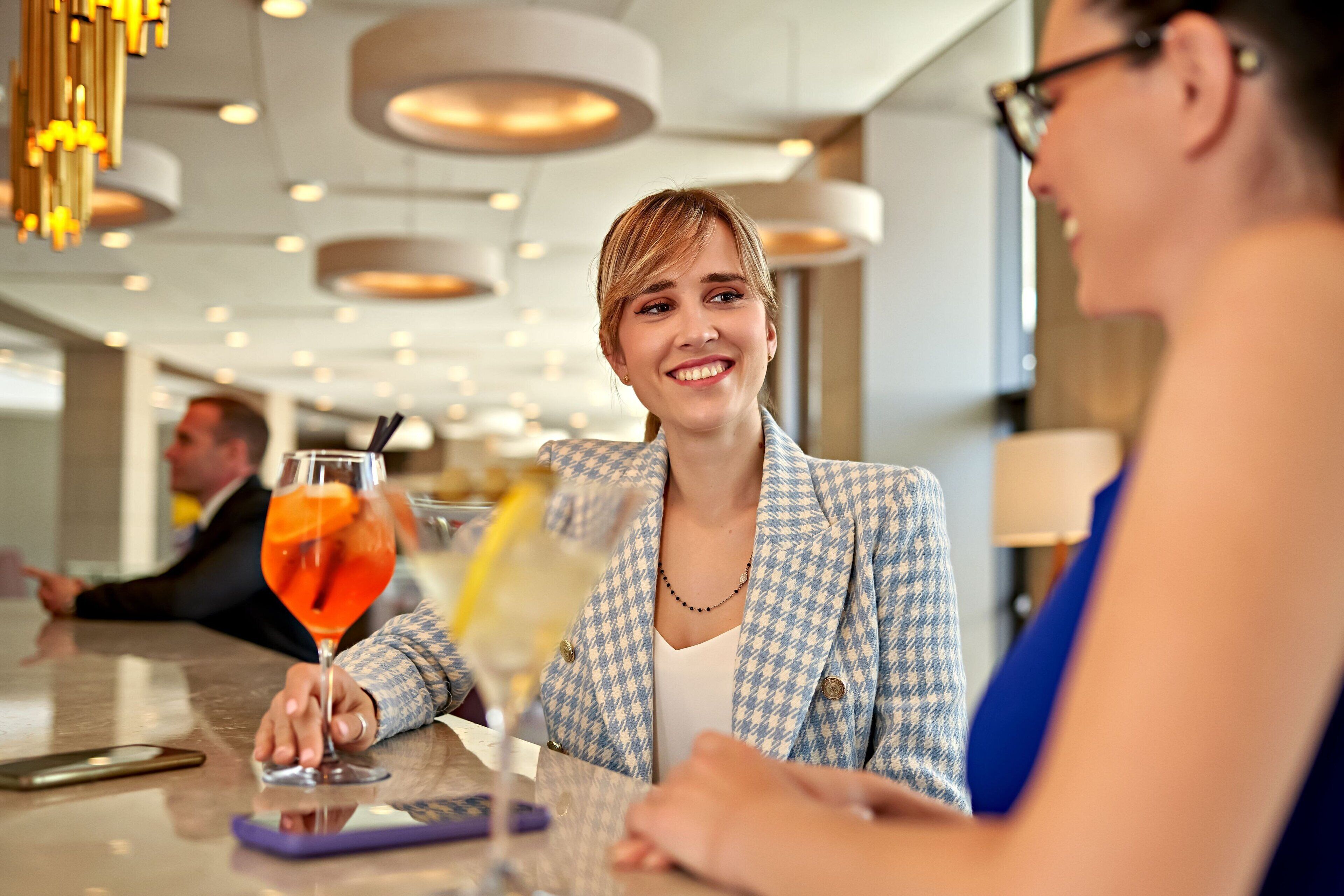 A person in a checkered blazer sitting at a table with a drink.
