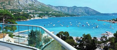 A coastal view from a balcony with a clear blue sea, boats, and mountainous terrain in the background.