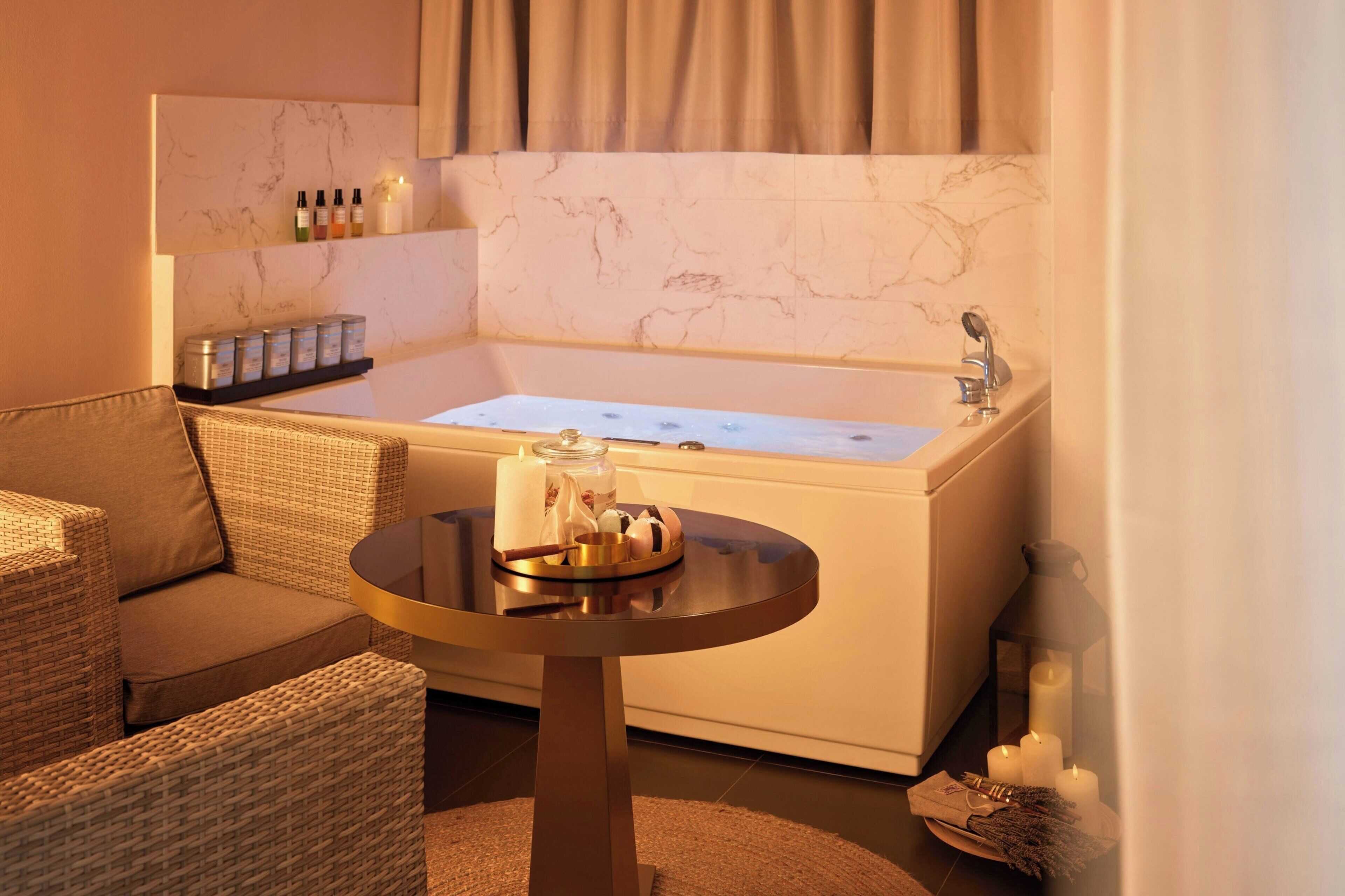 A bathroom with a bathtub, a small round table with toiletries, and a wicker chair.