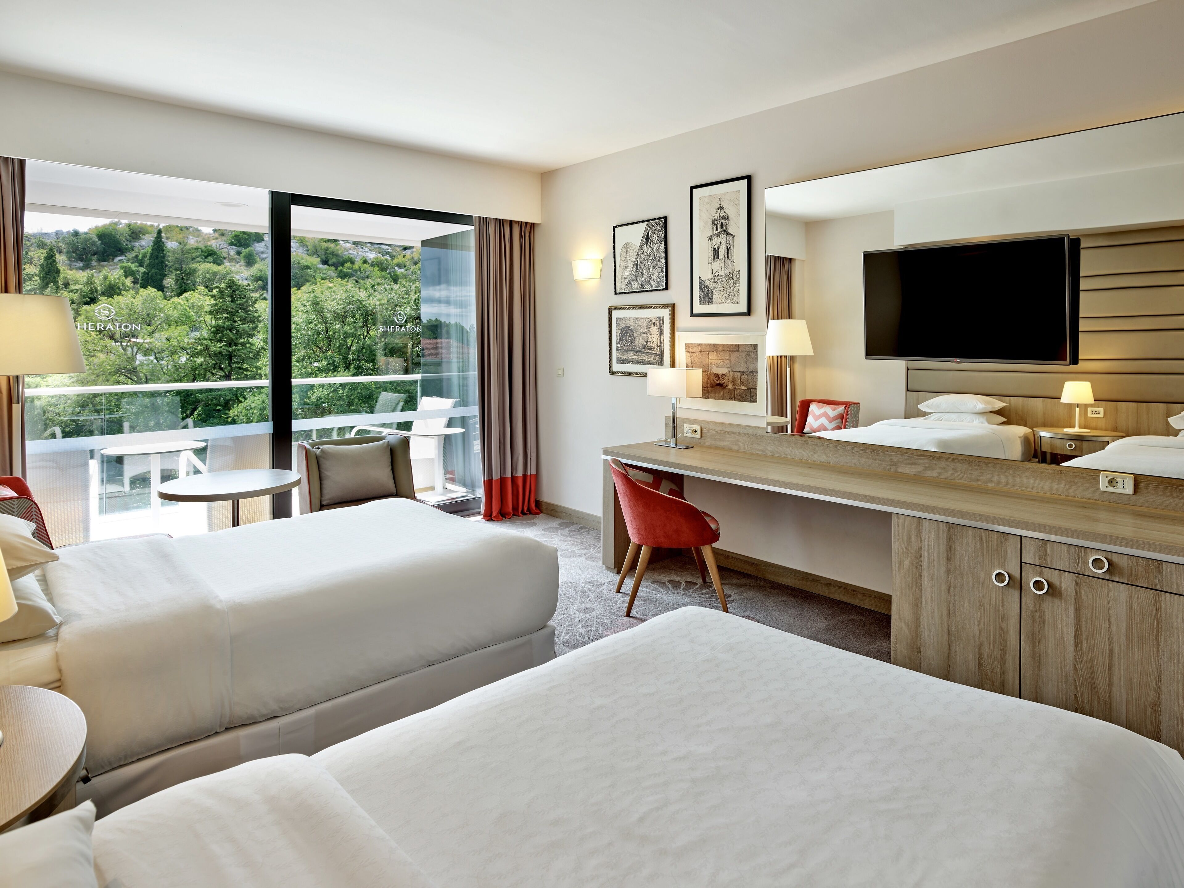 A hotel room with two beds, a desk, a TV, and a balcony with a view of greenery.