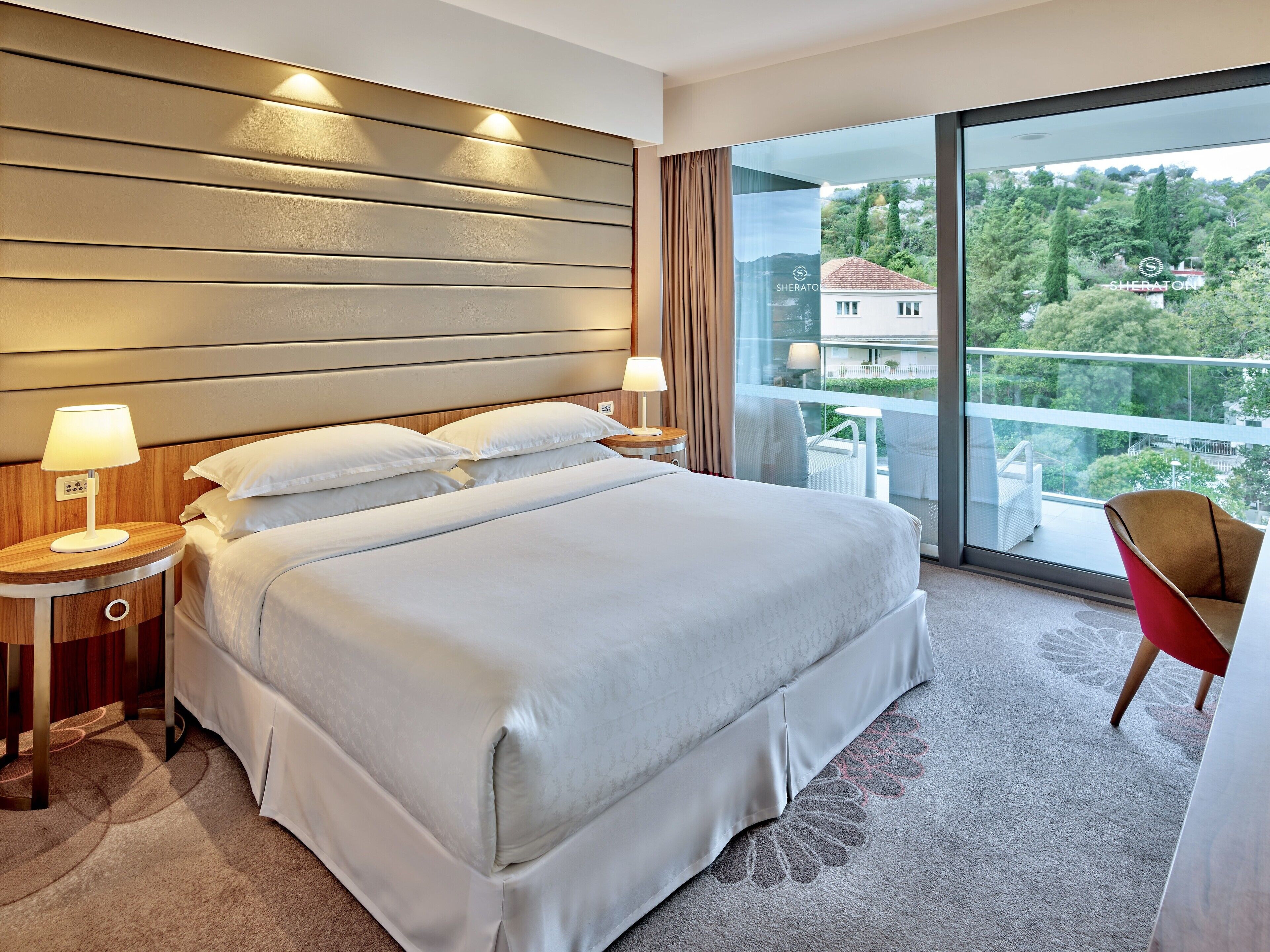 A hotel room with a large bed, two bedside tables with lamps, a red chair, and a balcony with a view of greenery and buildings.
