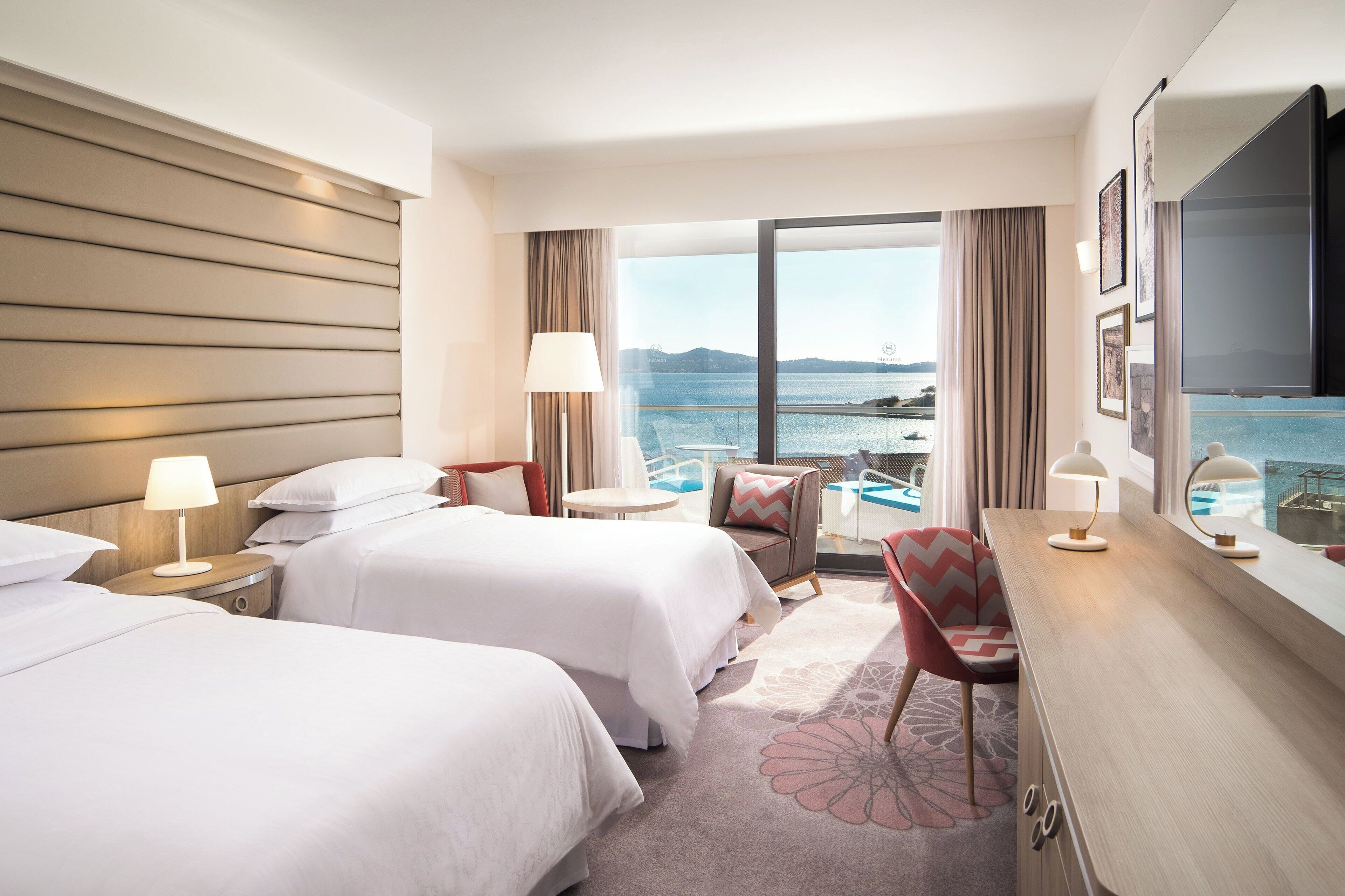 Habitación de hotel con dos camas, un escritorio, una silla, una mesita y un ventanal con vistas al mar.