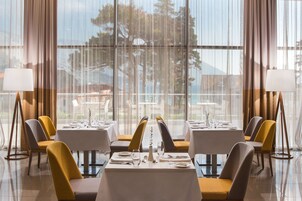 A dining area with tables set for four, chairs in yellow and grey, and a view of trees through large windows.