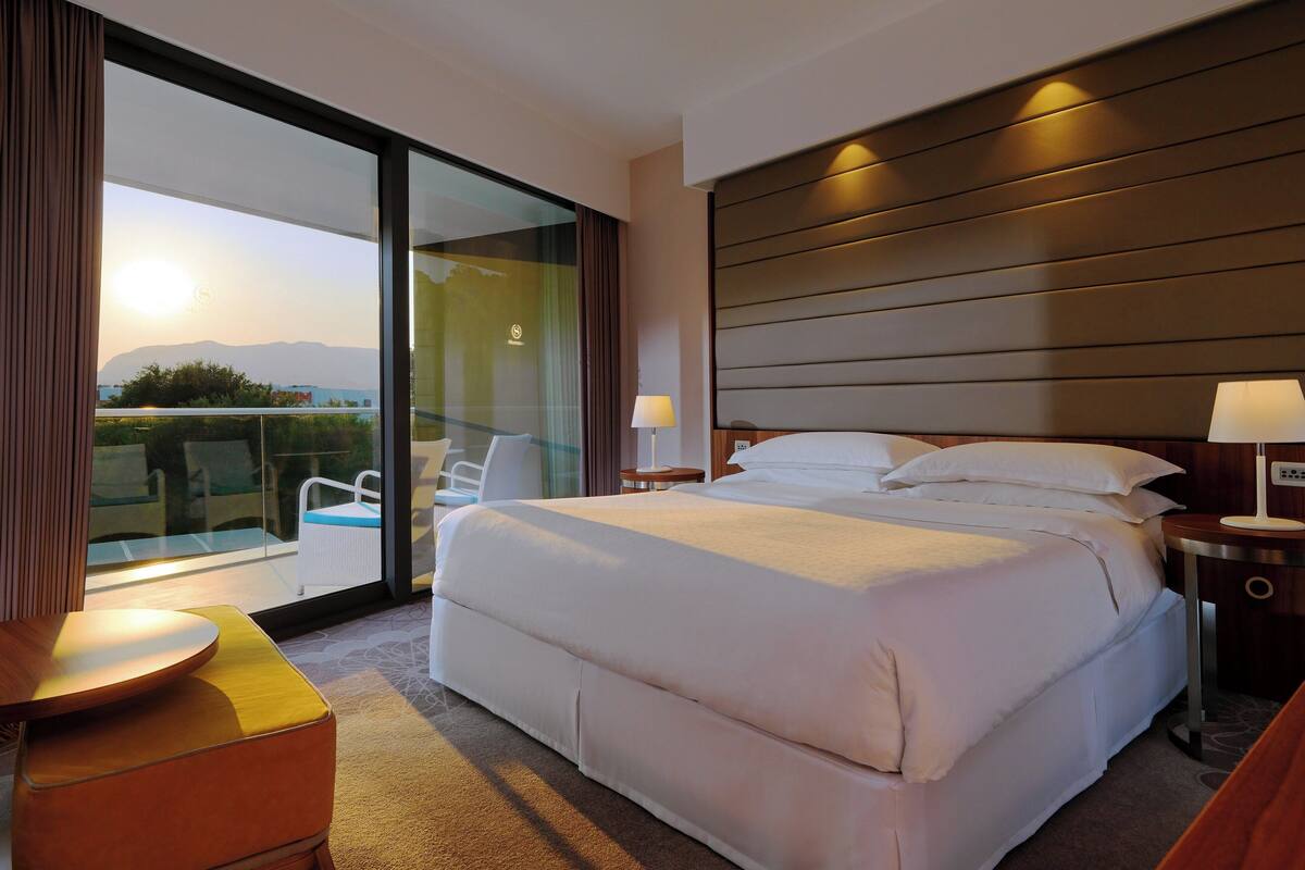 Une chambre d’hôtel moderne avec un grand lit, une porte vitrée donnant sur un balcon et une vue sur les montagnes.