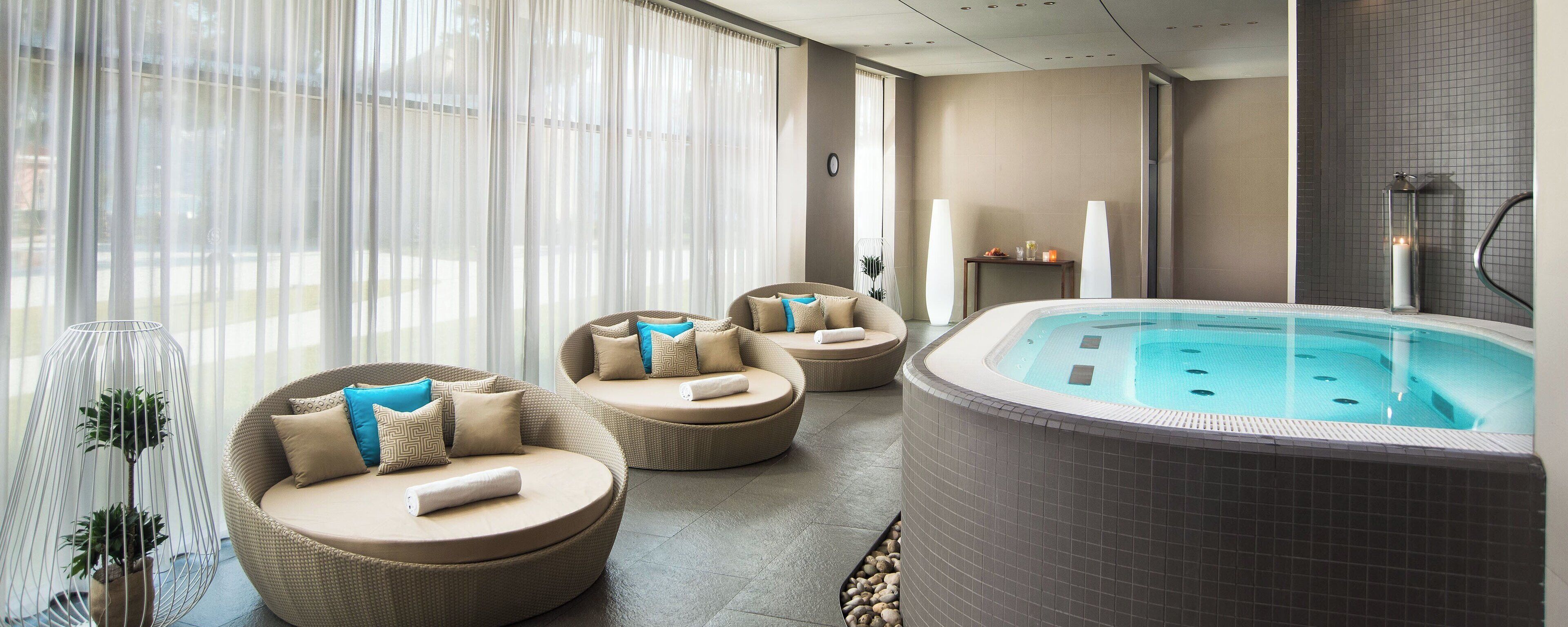 Un espace bien-être comprenant un jacuzzi, des canapés en osier avec des coussins et une grande fenêtre agrémentée de rideaux légers.