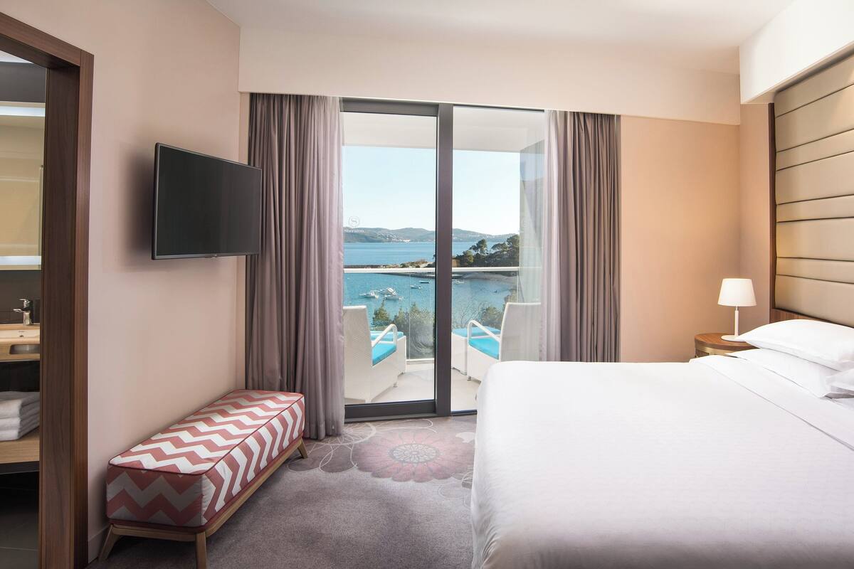 Une chambre d’hôtel avec un lit, une télévision, un banc et une vue sur une piscine et des montagnes.