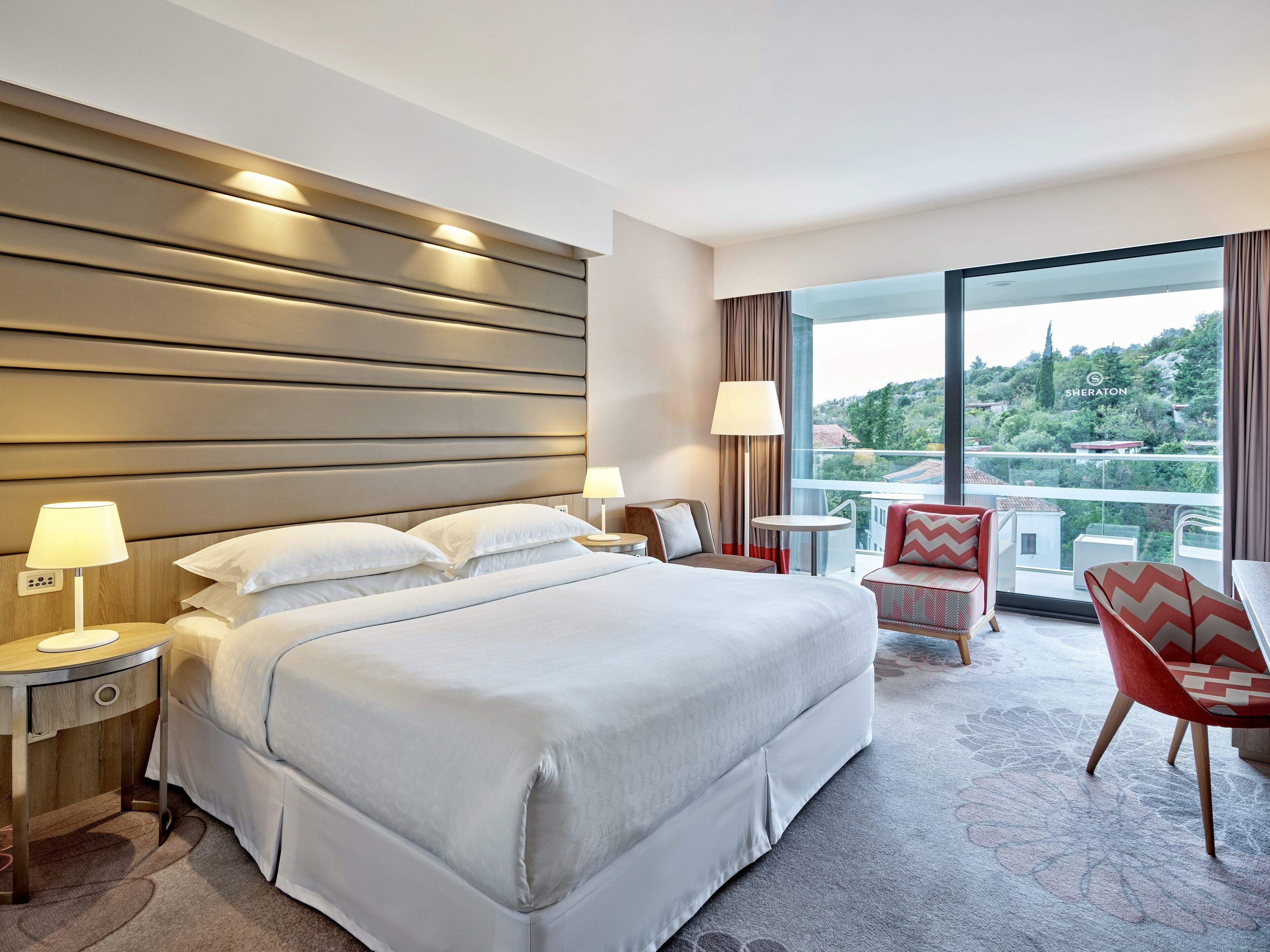 Une chambre d’hôtel avec un grand lit, deux tables de chevet avec des lampes, une petite table ronde, une chaise rouge et une grande fenêtre donnant sur la ville.
