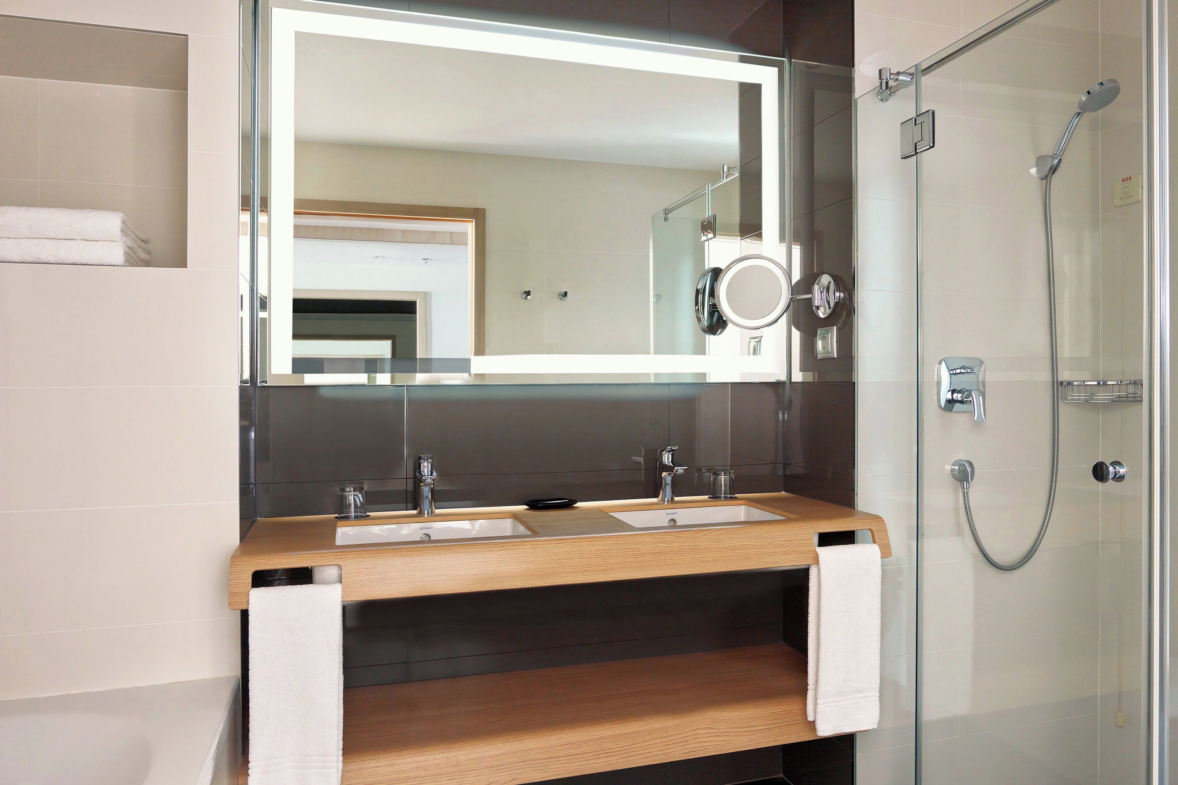 A modern bathroom with a wooden vanity, two sinks, a large mirror, and a glass-enclosed shower.