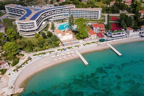 Vue aérienne d’un hôtel situé sur la côte, avec un bâtiment aux lignes courbes, une piscine et une plage aménagée avec des parasols.