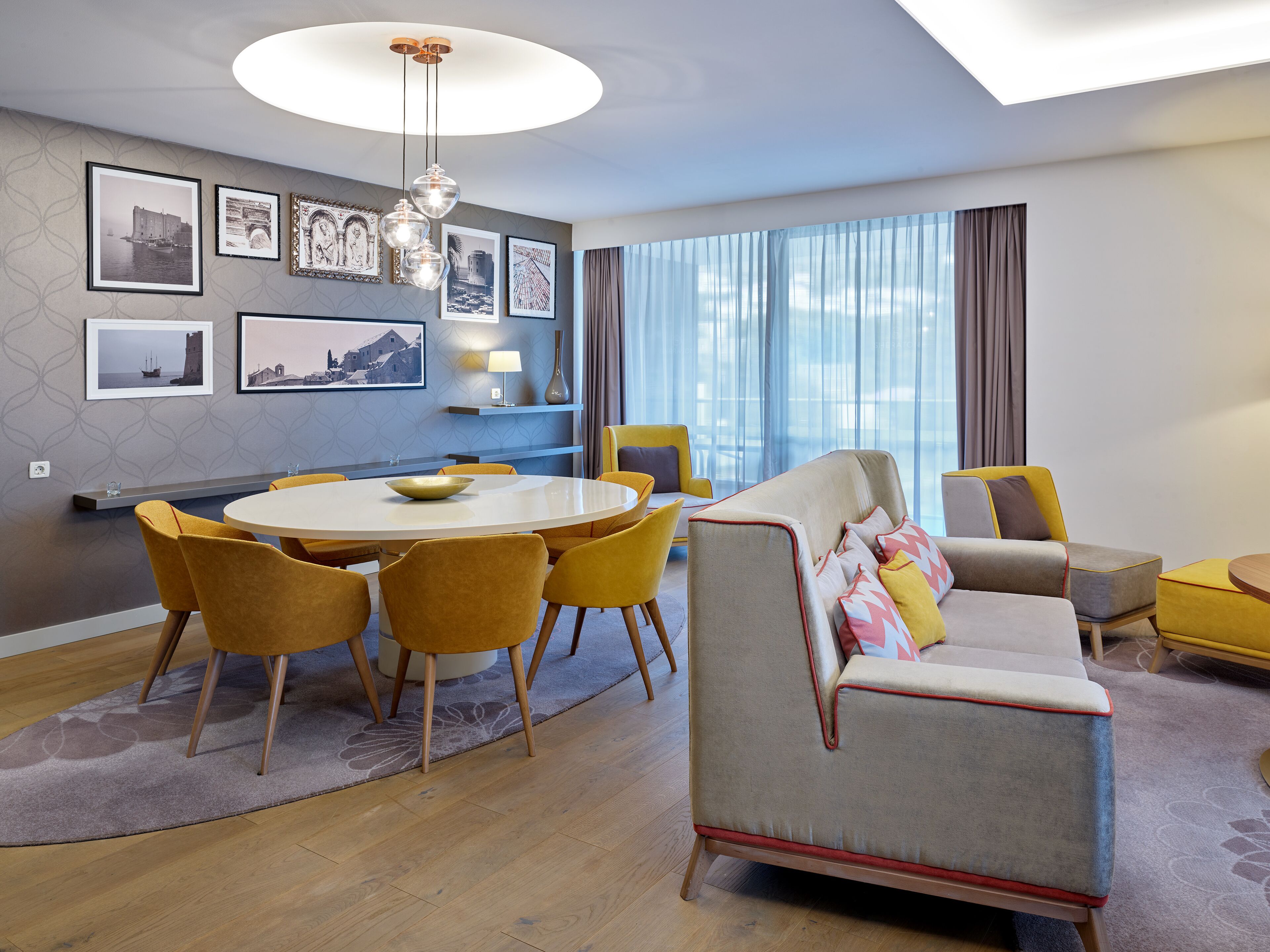 A modern living room with a round dining table, yellow chairs, a grey sofa with colorful cushions, and a large window with curtains.