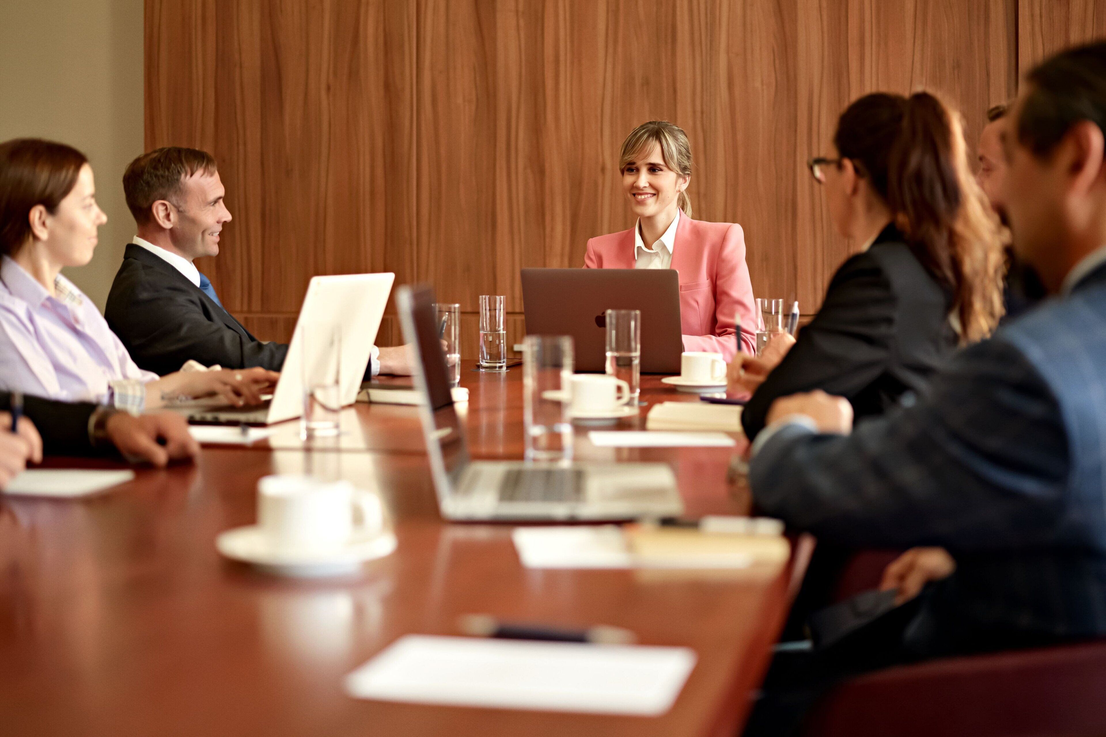 A conference room with a wooden paneled wall, a long table with laptops, cups, and documents.