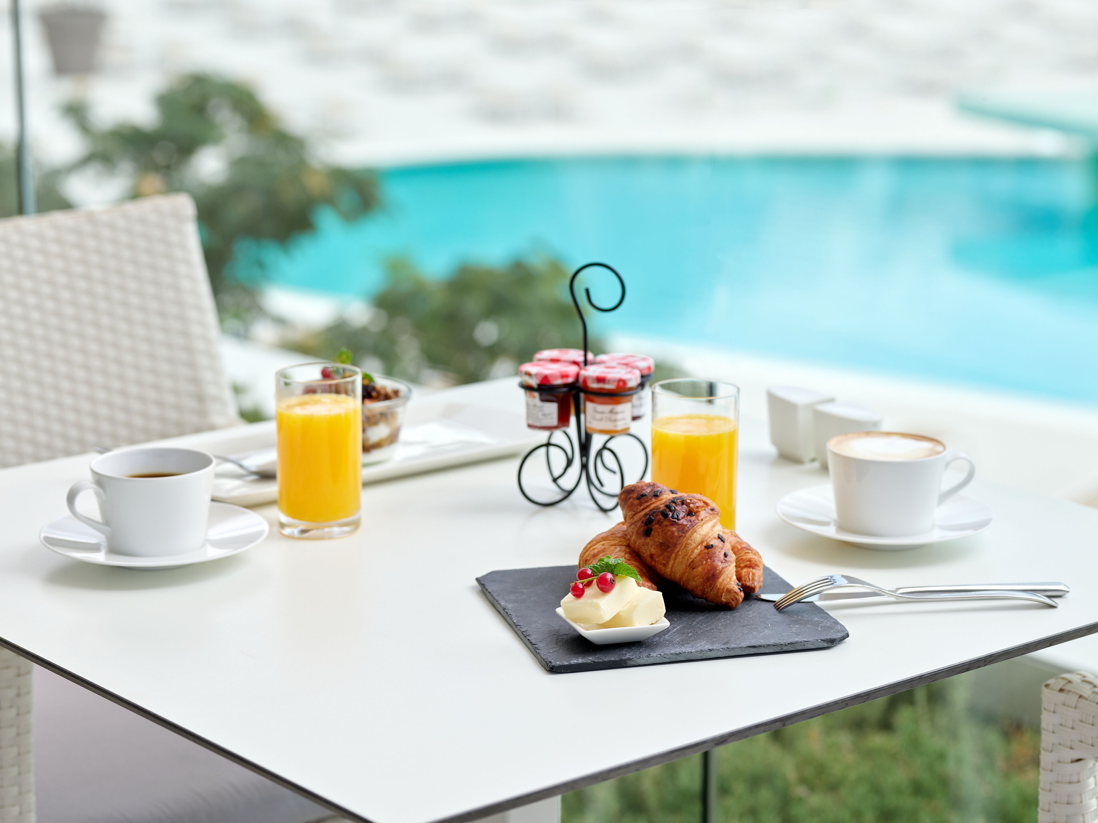 A breakfast setup with croissants, butter, and jam on a table by a poolside.