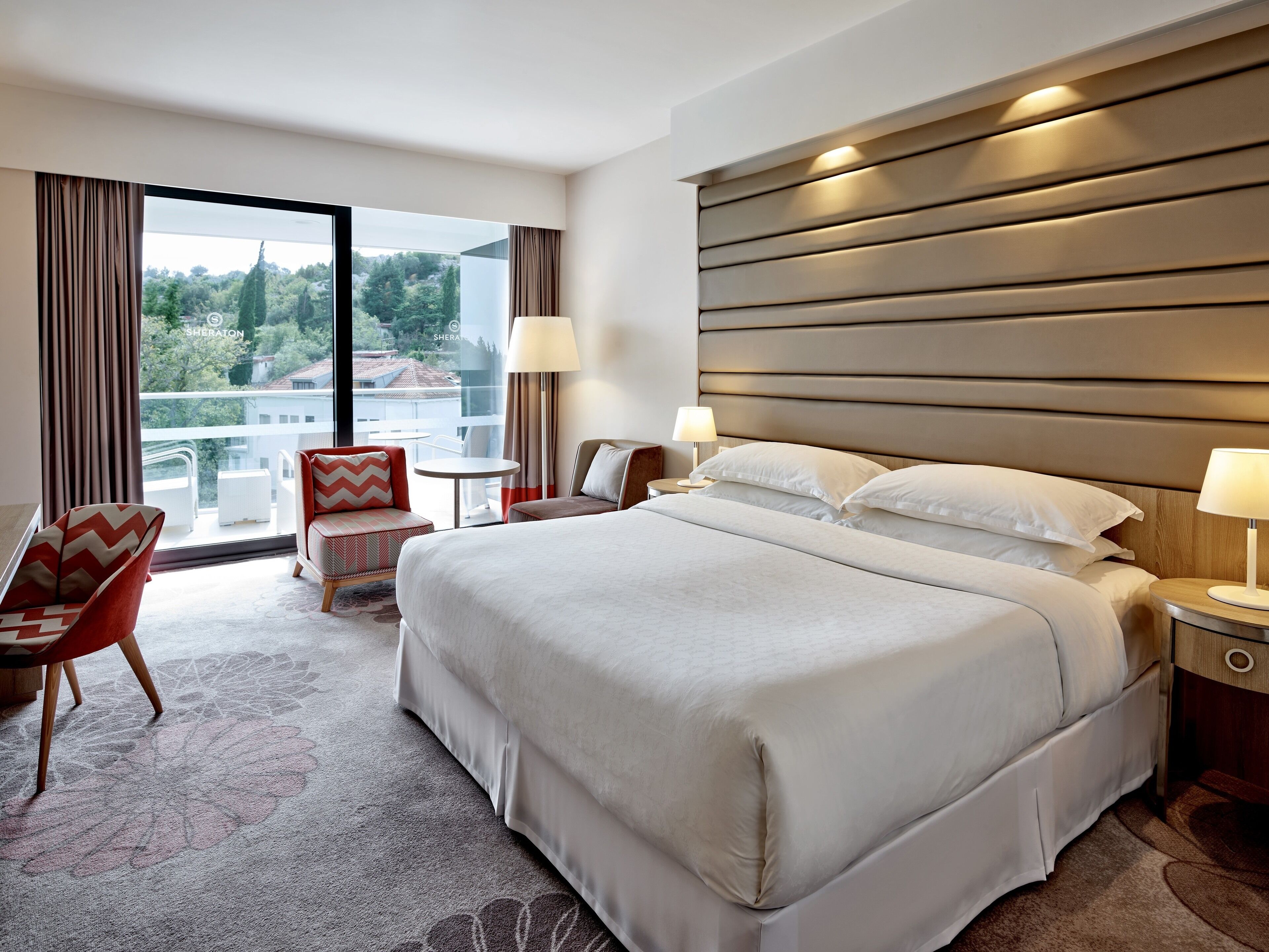 A hotel room with a large bed, two bedside tables with lamps, a chair, and a view of the outdoors.