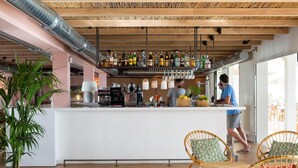 A modern bar area with a white countertop, various bottles, and a person working behind the bar. There are wicker chairs and a large potted plant in the foreground. - Hostal LA Savina (Formentera)