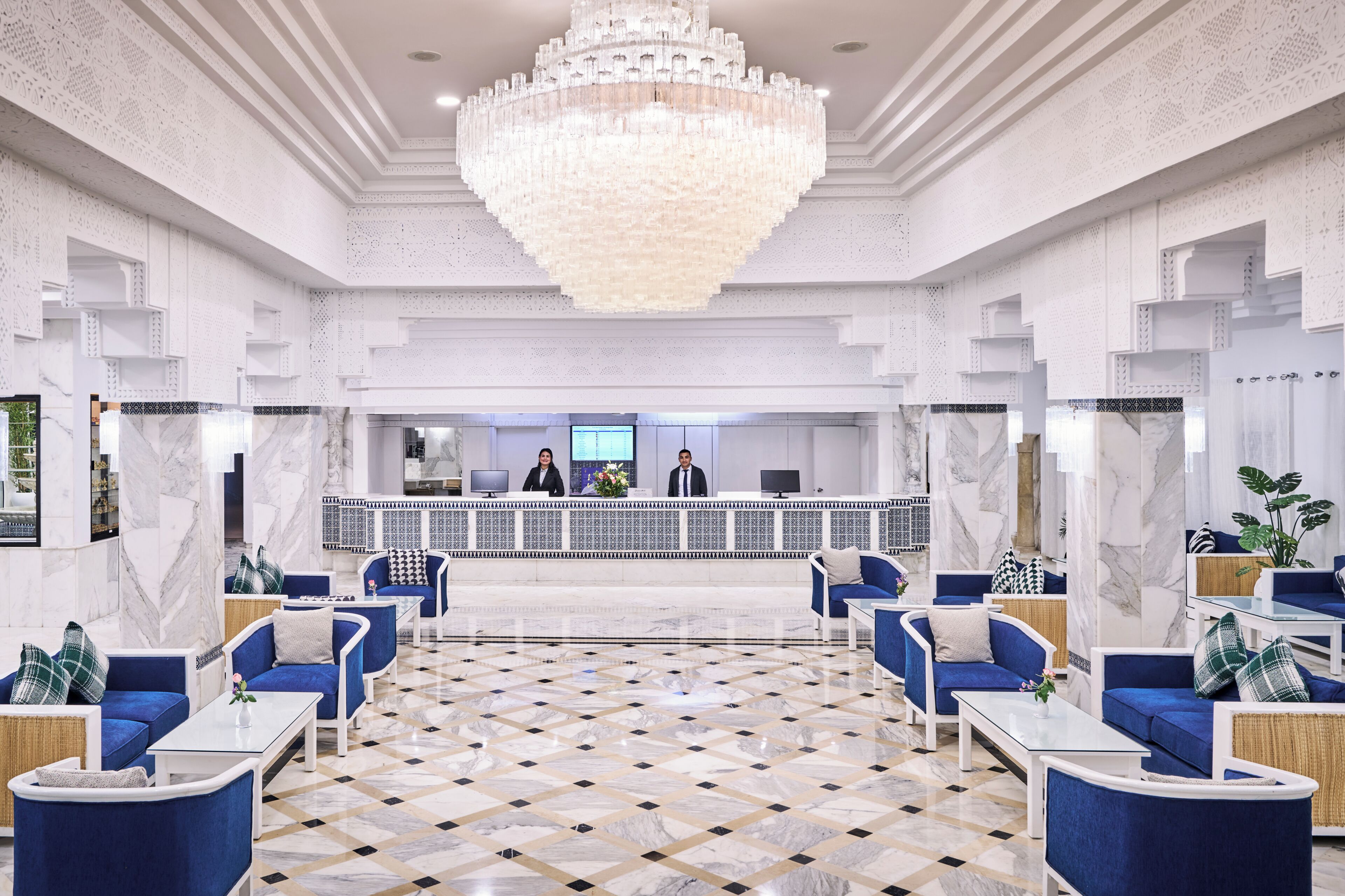 A spacious lobby with a marble floor, blue seating, a reception desk, and a large chandelier.