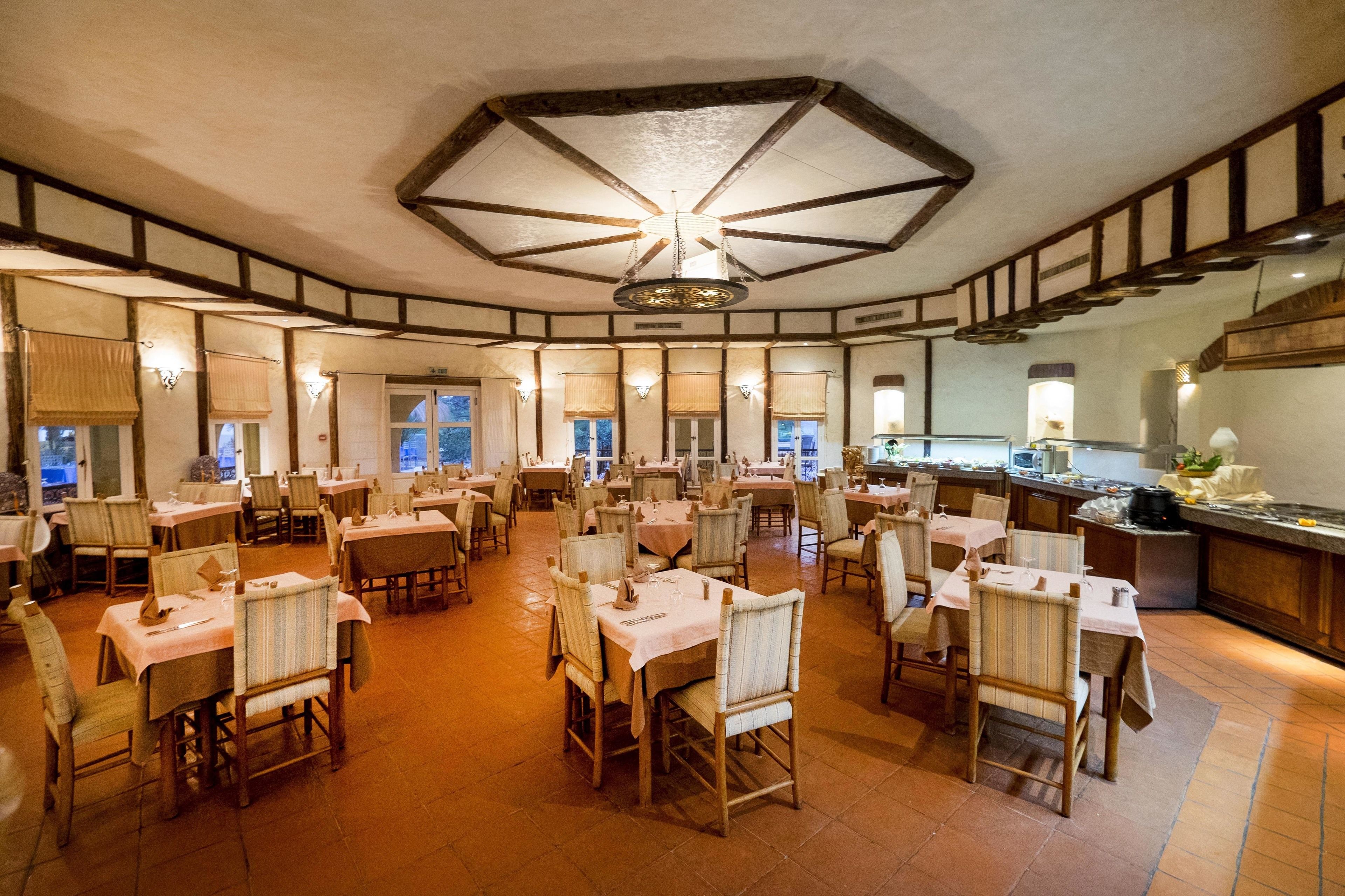 A spacious dining area with multiple tables and chairs, a central chandelier, and a kitchen area visible to the right.