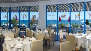 A restaurant with tables set for dining, large windows offering a view of the sea, and flags outside. - Primasol Sineva (Sveti Vlas)