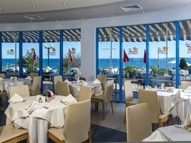Restaurante com mesas preparadas para refeições, amplas janelas com vista para o mar e bandeiras do lado de fora.