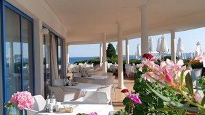 An outdoor dining area with tables and chairs, a view of the sea, and a building with large windows. - Primasol Sineva (Sveti Vlas)