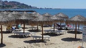 A beach with thatched umbrellas and lounge chairs, a clear sky, and a distant view of a cityscape and bridge. - Primasol Sineva (Sveti Vlas)
