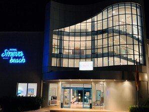 Hotel com fachada moderna em vidro, escadaria visível e letreiro luminoso indicando o nome 'Sineva Beach'.
