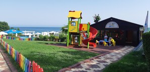 A playground with a slide, surrounded by a garden and a building labeled "Prima Kids Club". - Primasol Sineva (Sveti Vlas)