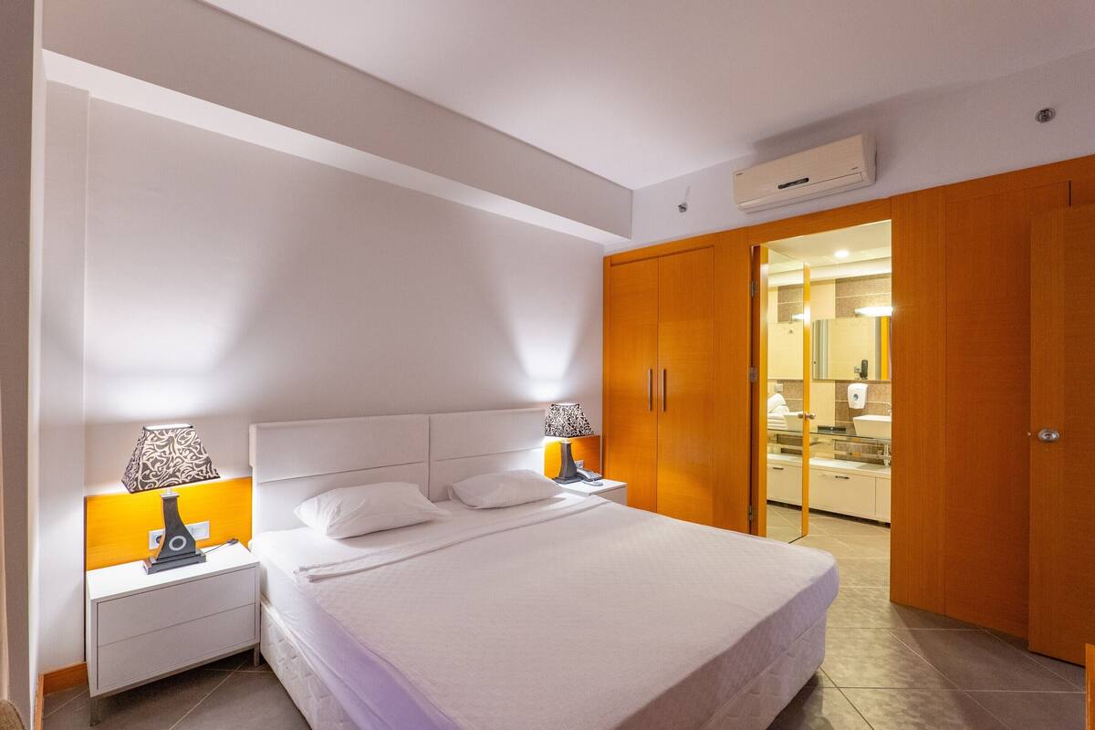A hotel room with a large bed, two bedside tables with lamps, a wardrobe, and a bathroom visible through an open door.