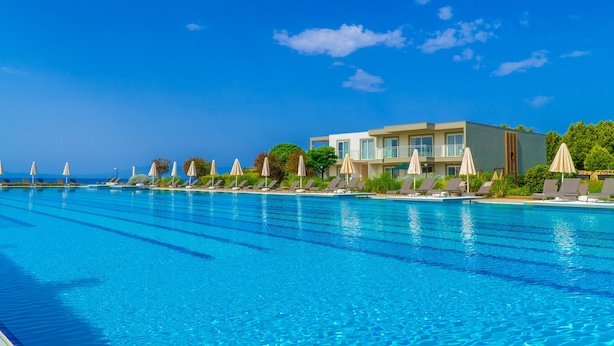 A swimming pool with lounge chairs and umbrellas, a building with large windows, and a clear blue sky. - Karya Family Resort – All inclusive (Menderes)
