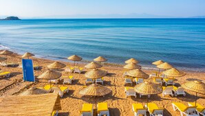 Une plage avec des chaises longues et des parasols en chaume, un panneau indiquant la direction de Karya, et une vue dégagée sur la mer et les montagnes lointaines.