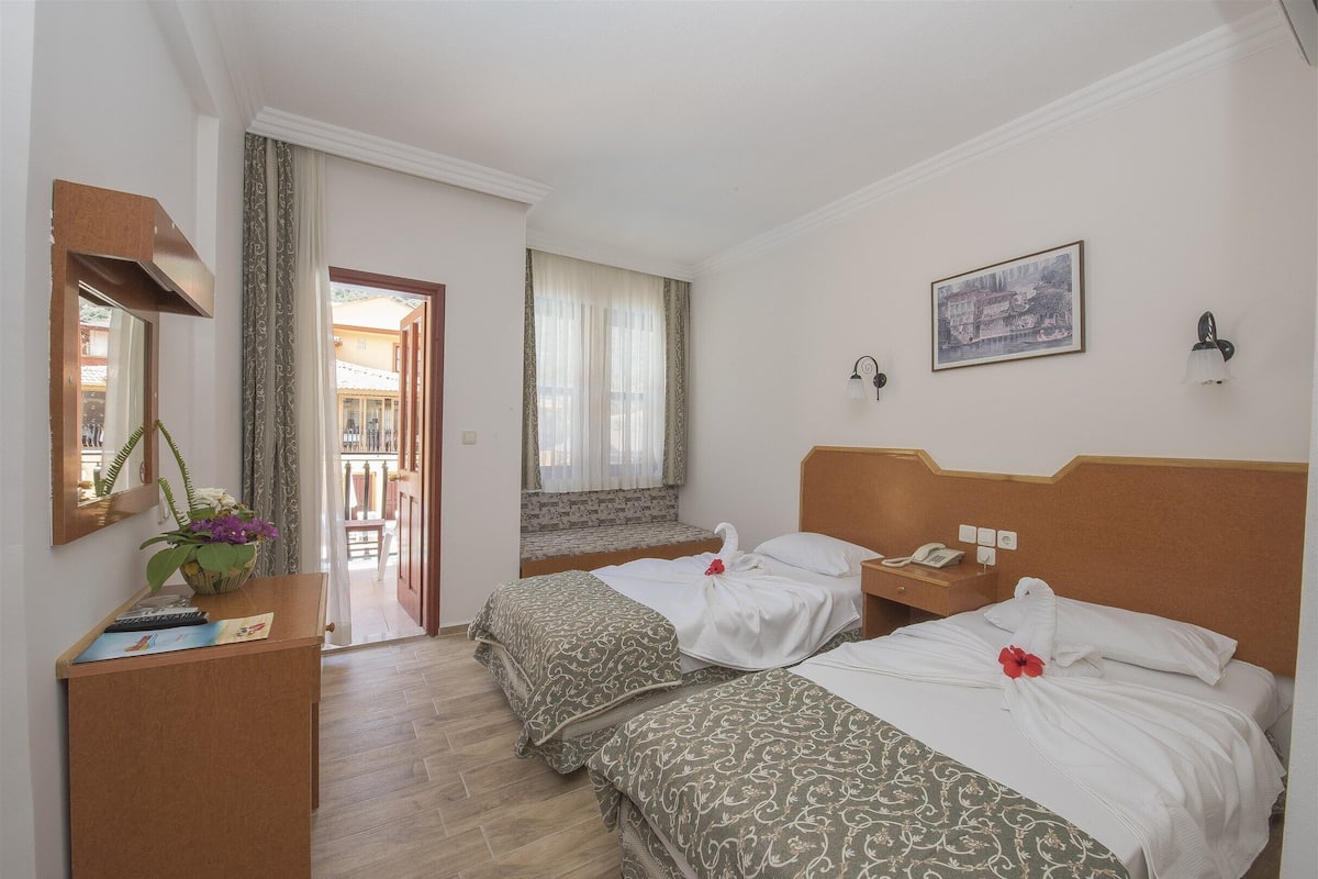 A hotel room with two beds, a desk, a mirror, a window with curtains, a painting on the wall, and a view of the outside balcony.