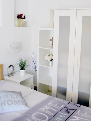 Un dormitorio con una cama, una mesita de noche con una planta, un armario y una estantería con objetos decorativos.