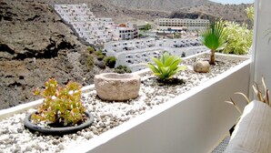 A balcony with a view of a coastal town, featuring white buildings, rocky terrain, and potted plants. - Apartamentos Malibu (Mogan)