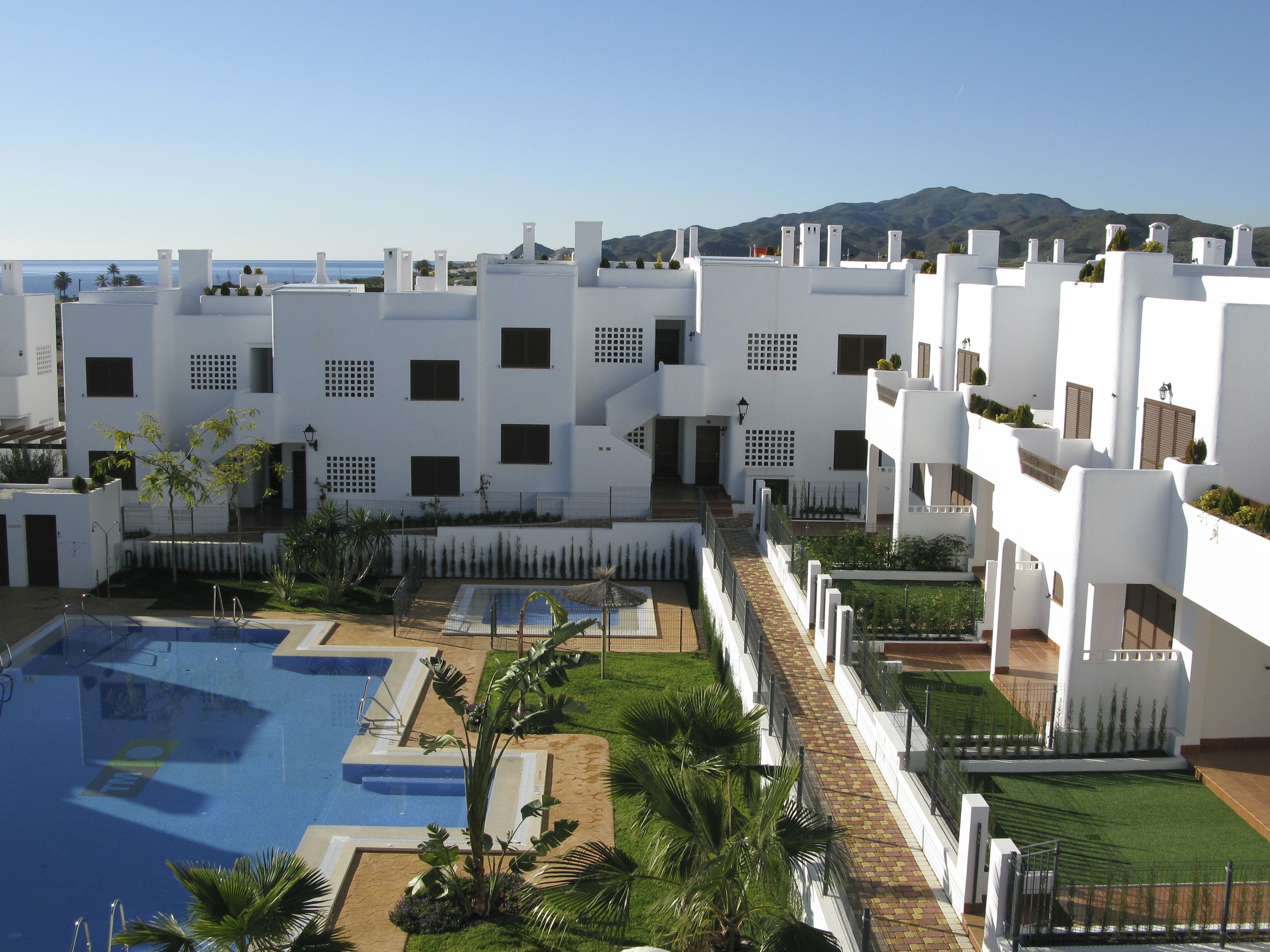 Un complejo turístico con varias edificaciones blancas, una piscina y vistas al mar y a las montañas.