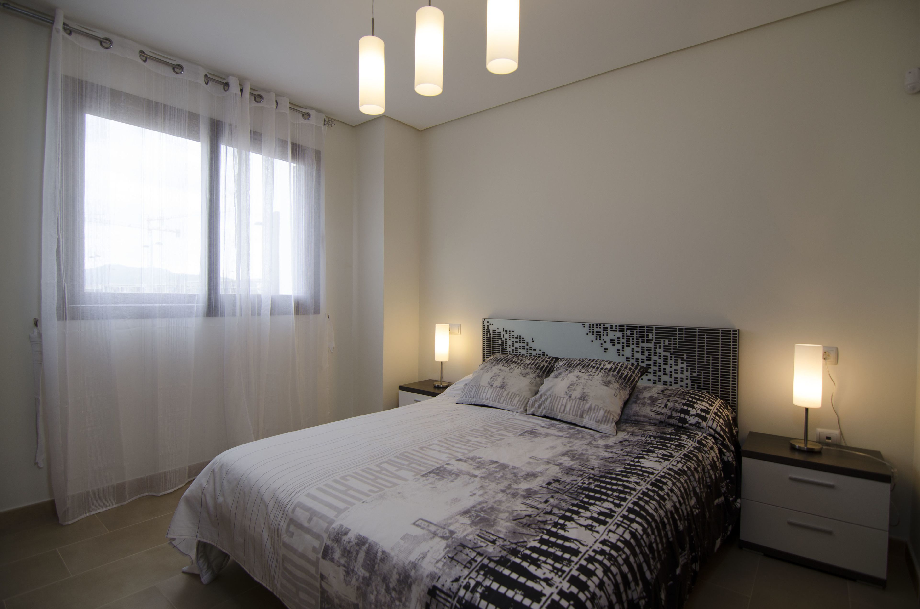 Un dormitorio con cama, dos mesitas de noche, una ventana con cortinas translúcidas y una lámpara en el techo.