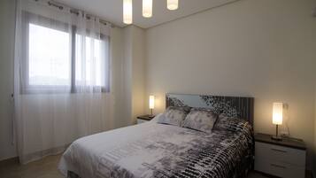 Un dormitorio con cama, dos mesitas de noche, una ventana con cortinas translúcidas y una lámpara en el techo.