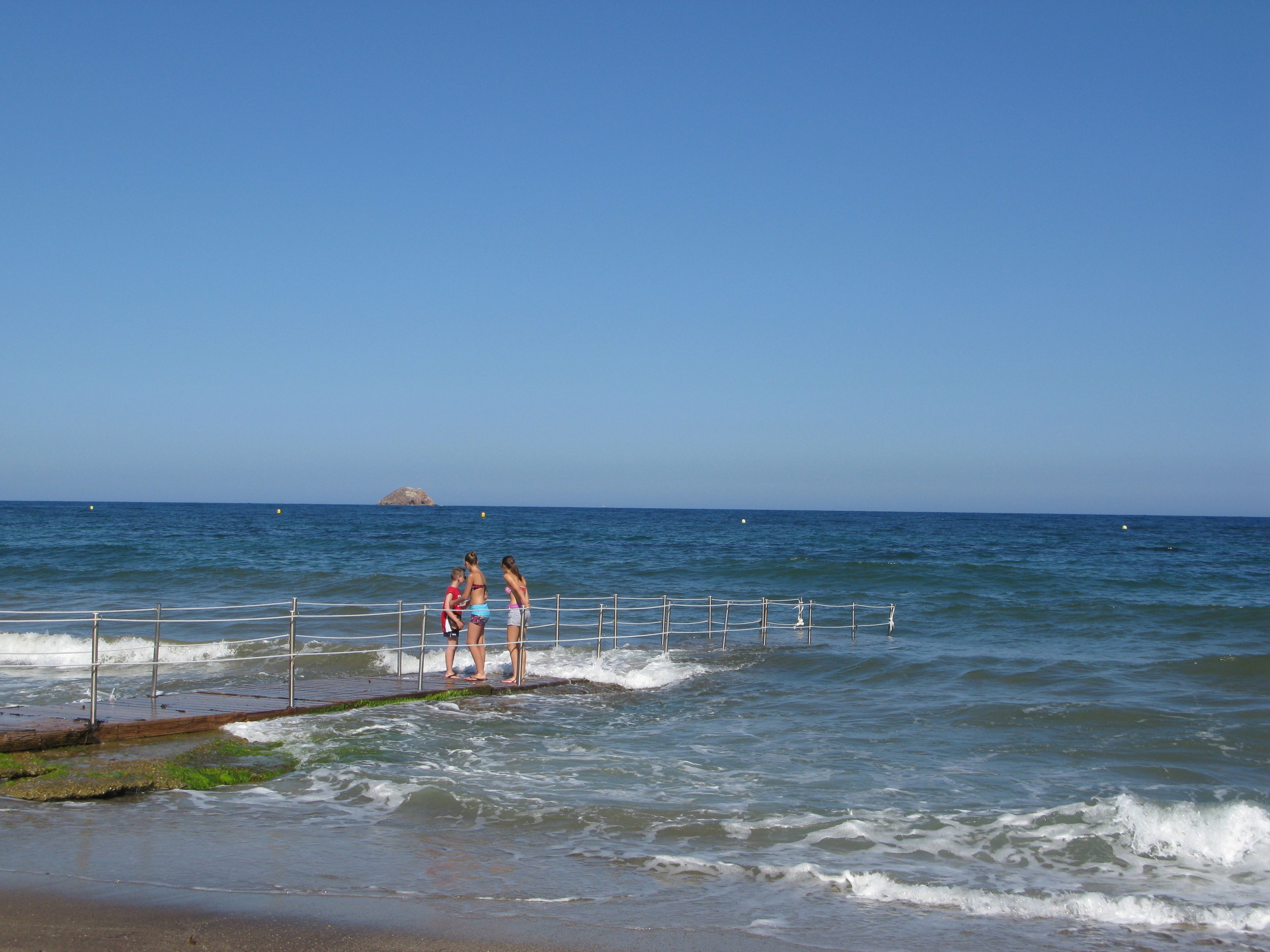 A beach with a wooden walkway leading to the water, a small island in the distance, and a clear blue sky.