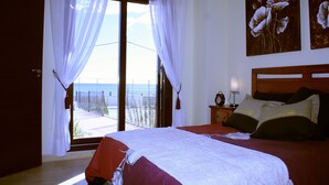 Une chambre avec un grand lit, une table de chevet avec une lampe et une vue sur la mer, visible à travers une fenêtre ornée de rideaux légers.