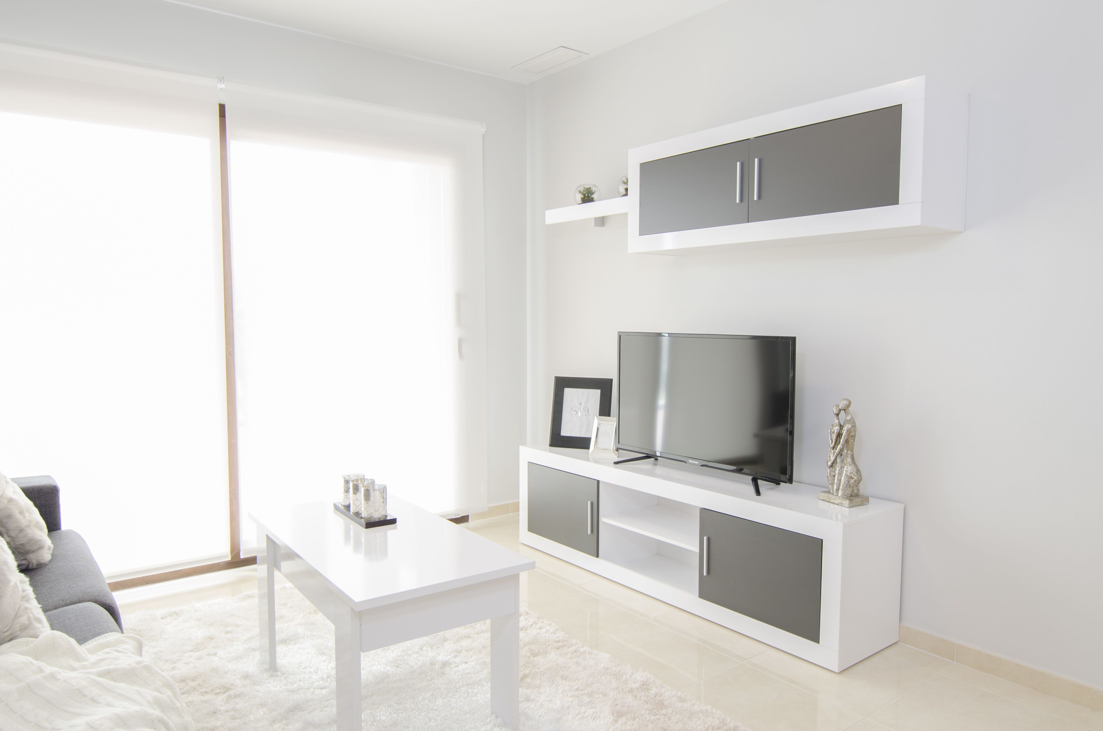 A modern living room with a white sofa, a white coffee table, a flat-screen TV on a white entertainment unit, and a large window providing natural light.