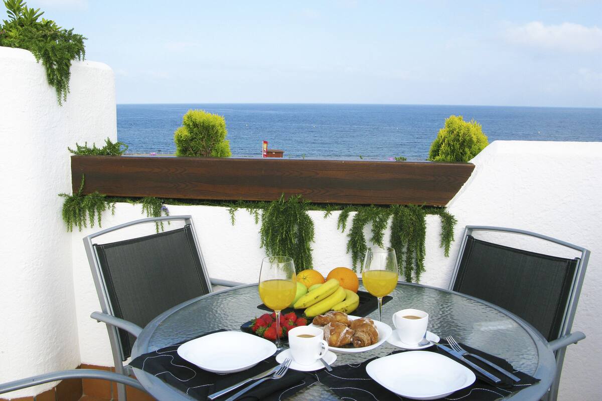 Une table dressée pour le petit-déjeuner, avec vue sur la mer, comprenant des fruits, du café et un verre de jus d’orange.