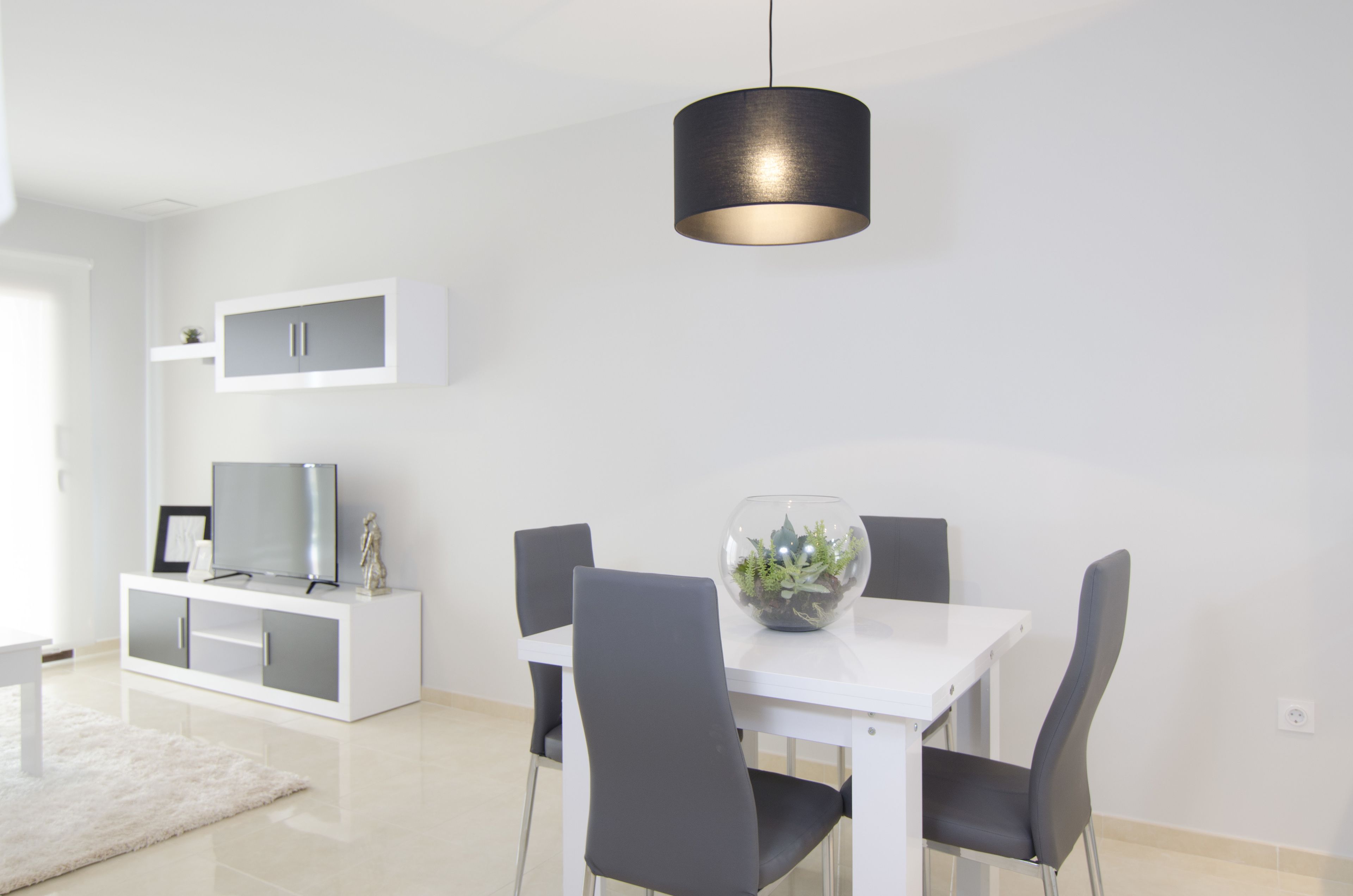 Un comedor moderno con una mesa blanca, cuatro sillas, un cuenco de vidrio con plantas, un televisor sobre un mueble blanco y una lámpara colgante negra.