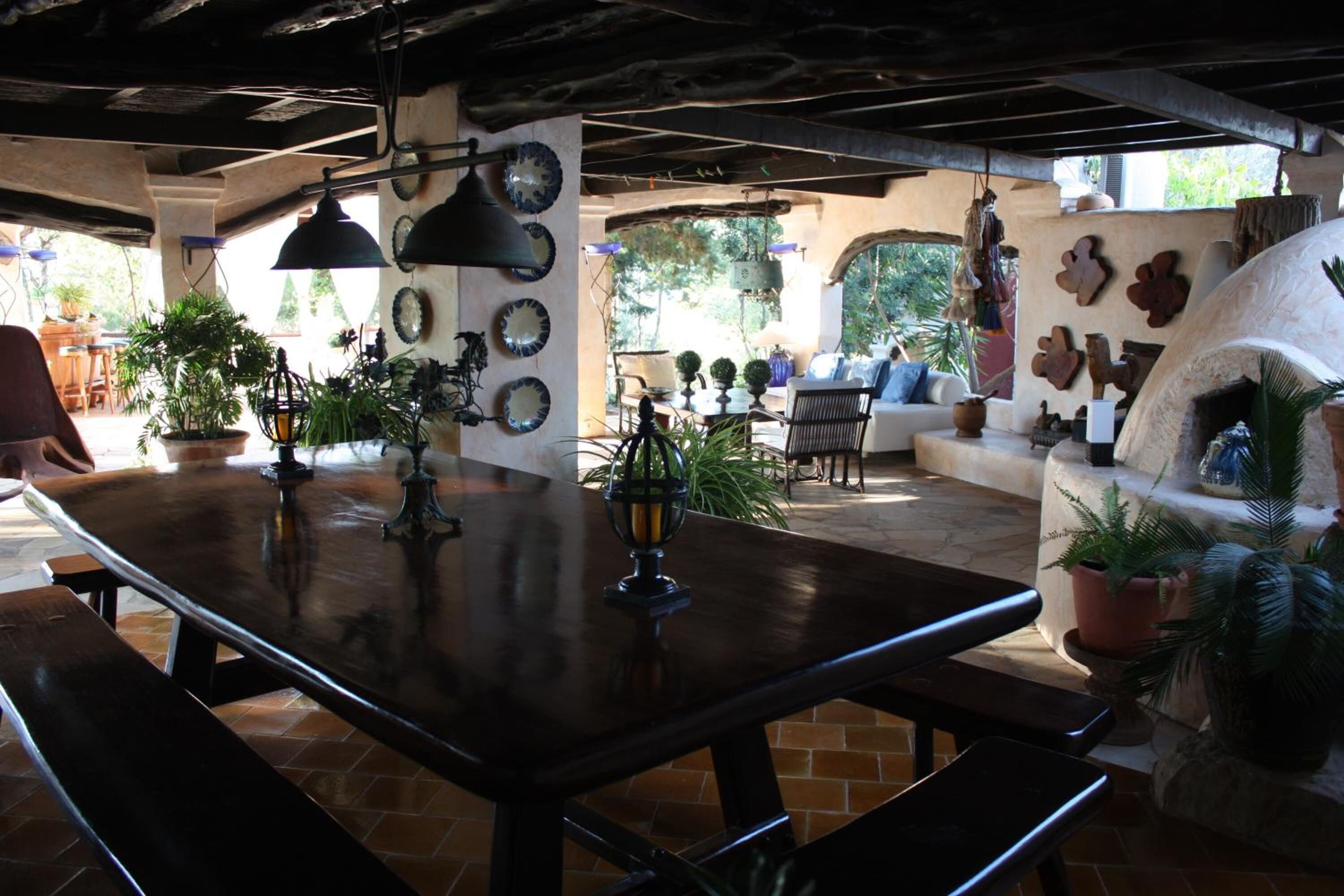 Un patio al aire libre con mesa de madera, bancos, plantas en macetas y un horno de piedra.