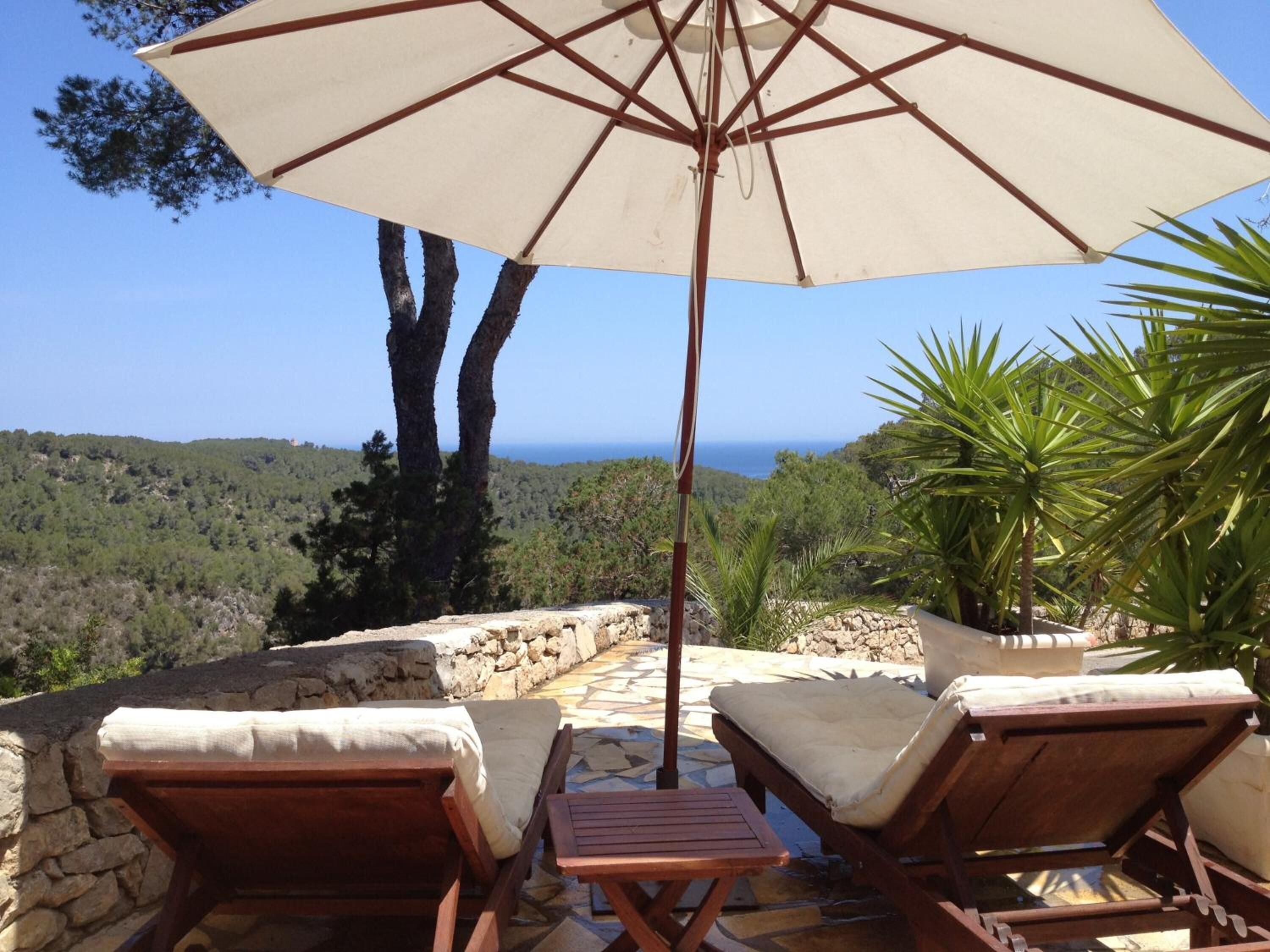 Une terrasse avec deux fauteuils de jardin en bois et une table, un parasol, et une vue sur la mer et la forêt.