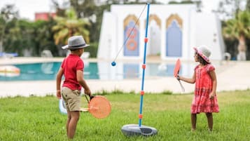 Children play badminton in a lush grassy area overlooking a swimming pool.