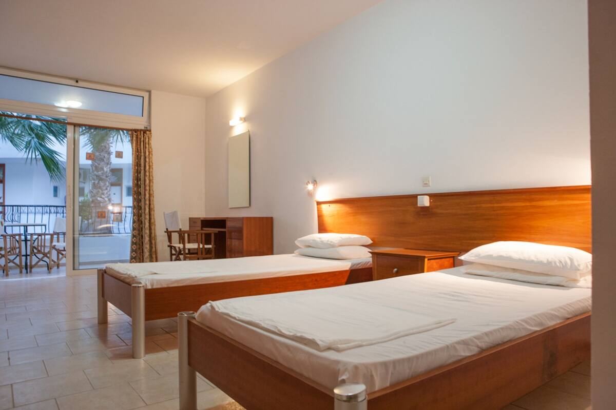 A hotel room with two single beds, a wooden headboard, a desk with a chair, and a view of a balcony with palm trees.