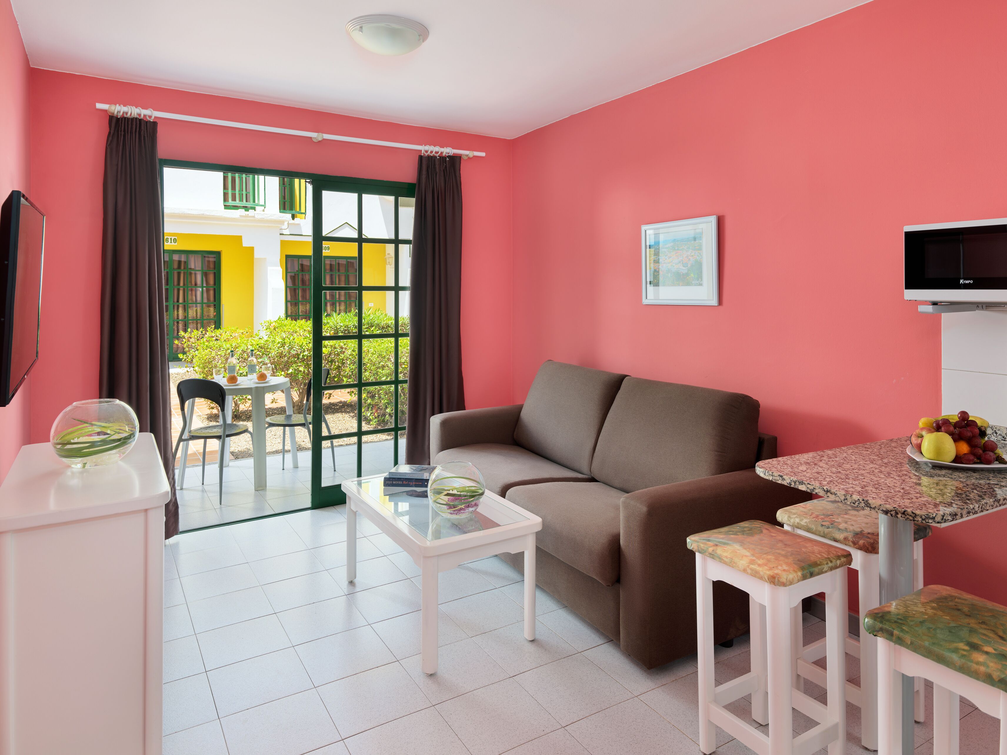 A room with a sofa, a table with fruit, a TV, and a view of a yellow building through a window.